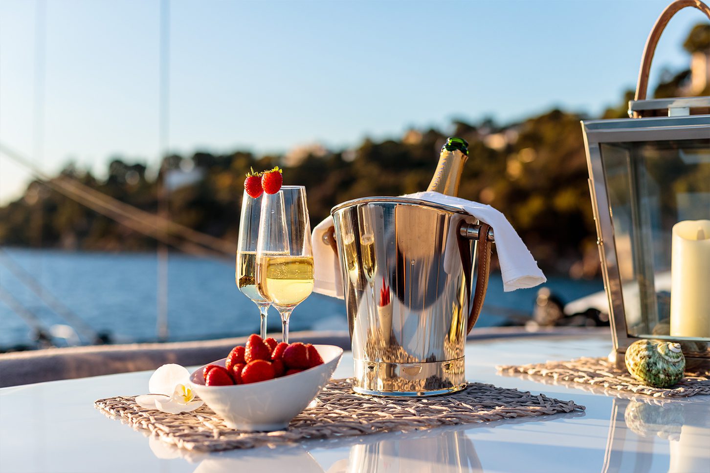Two champagne glasses with strawberries, a chilled bottle in an ice bucket, and a bowl of fresh strawberries on a yacht table at sunset.