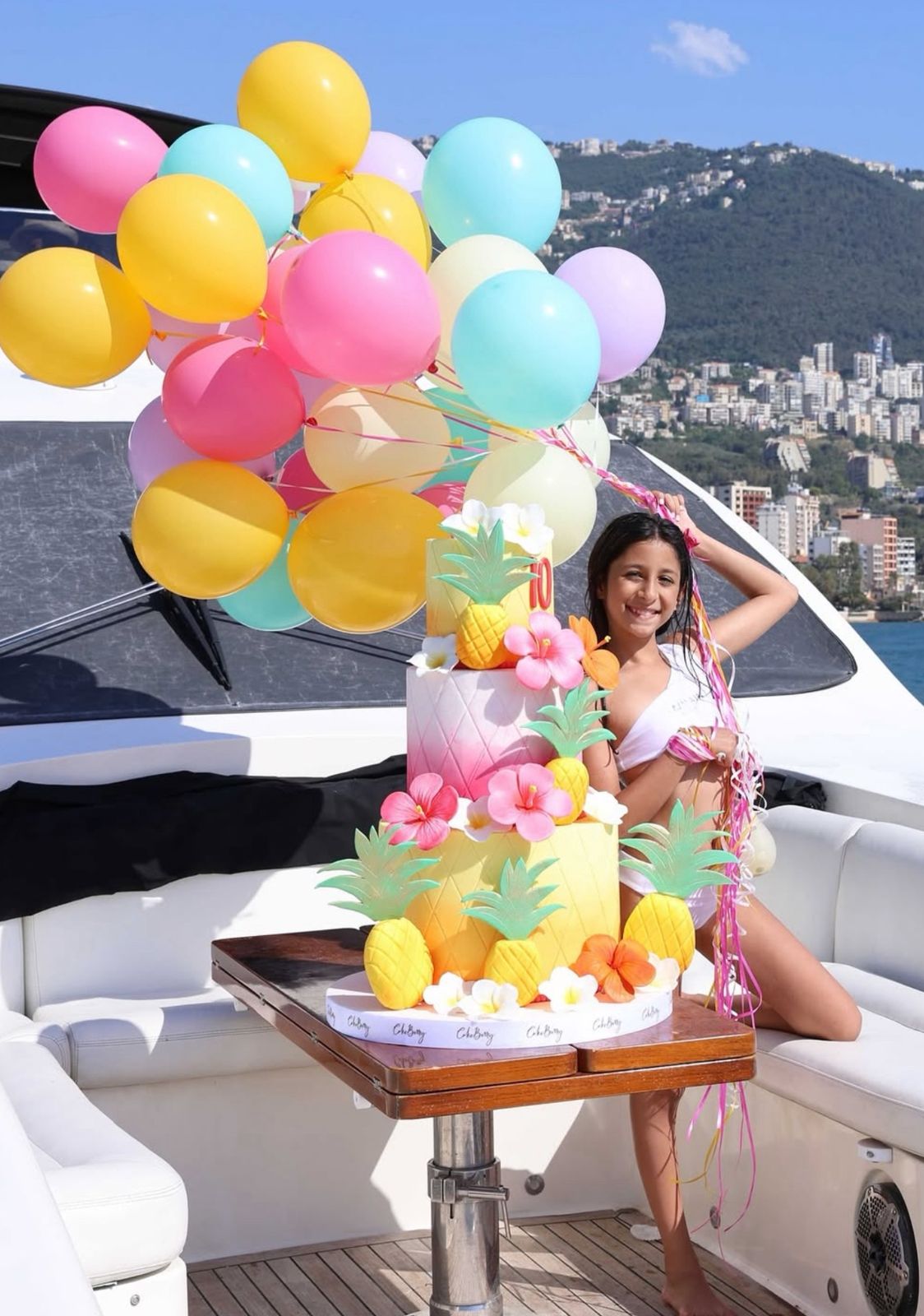 Smiling girl celebrating her birthday on a luxury yacht in Lebanon, with colorful balloons and a tropical pineapple-themed cake.
