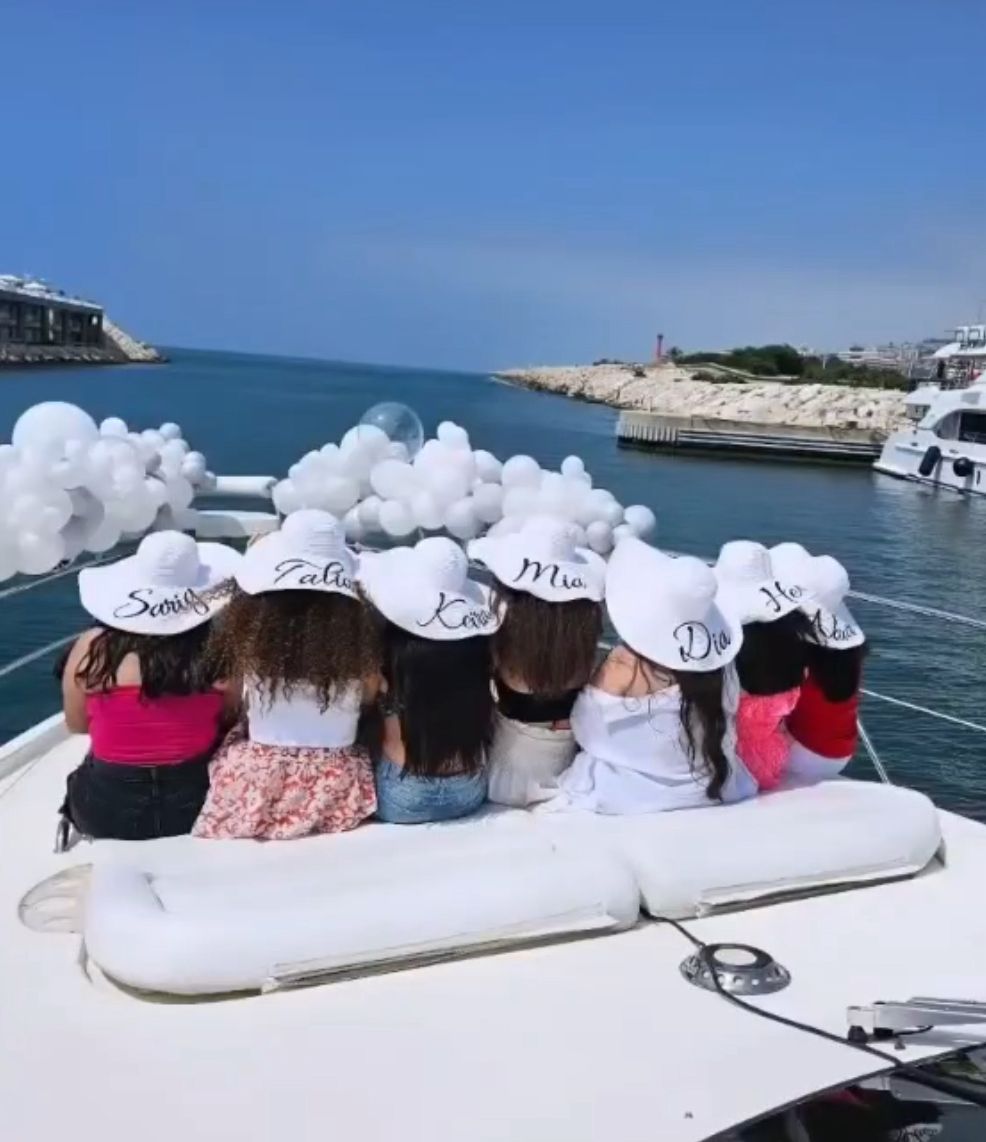 Group of girls wearing personalized white hats sitting together on a luxury yacht decorated with white balloons in Lebanon, celebrating under a bright blue sky.