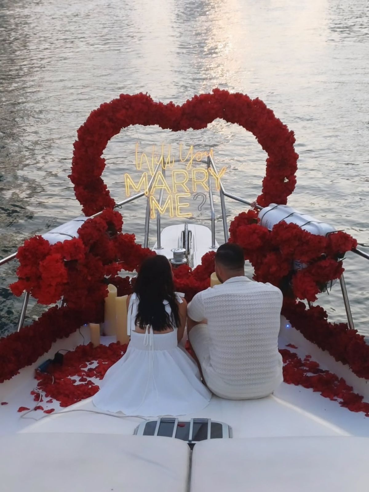 Couple seated in front of a red rose heart arch with a Will You Marry Me neon sign on a luxury yacht at sunset in Lebanon