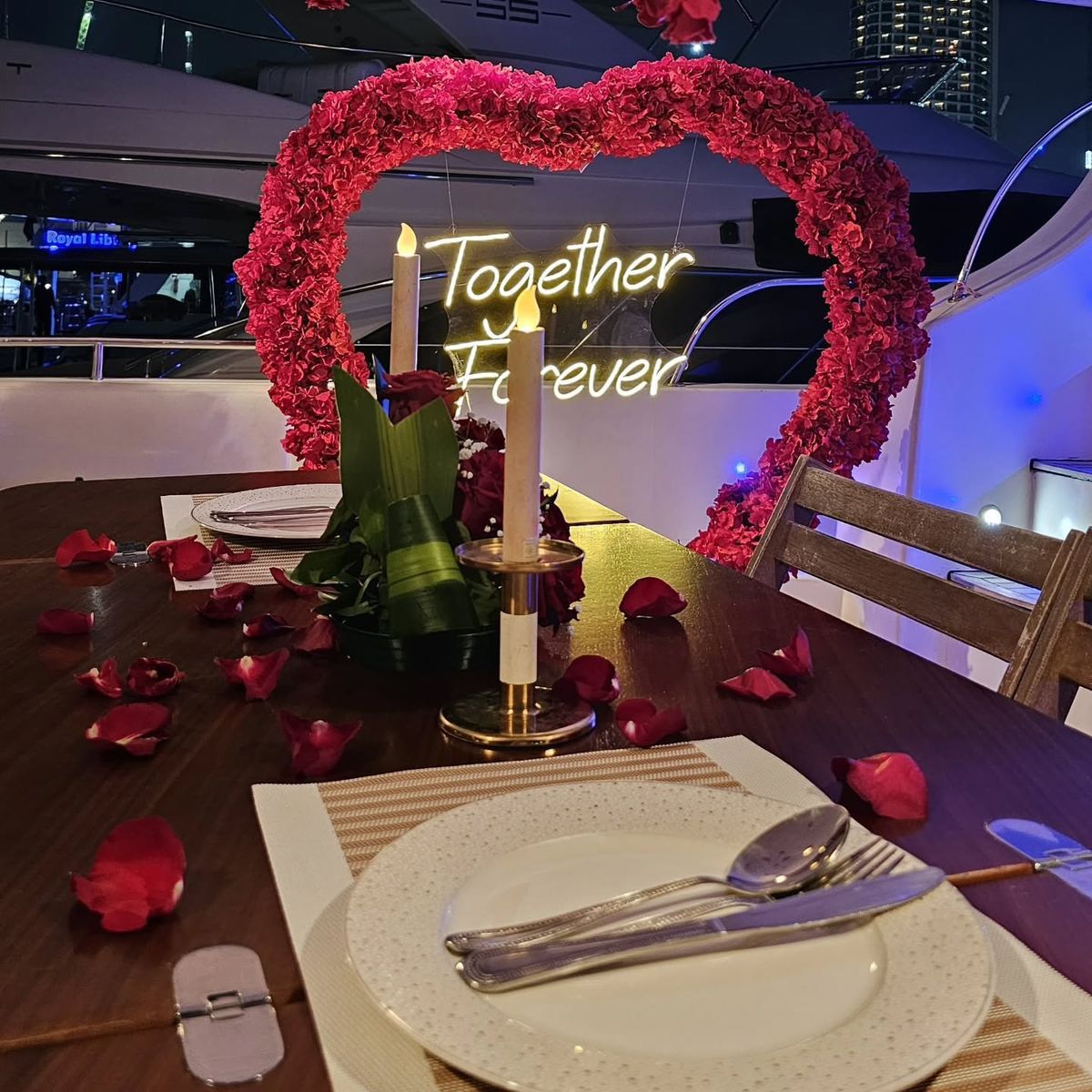 Romantic candlelit dinner setup on a luxury yacht deck with a rose heart arch and Together Forever neon sign, rose petals scattered across the table